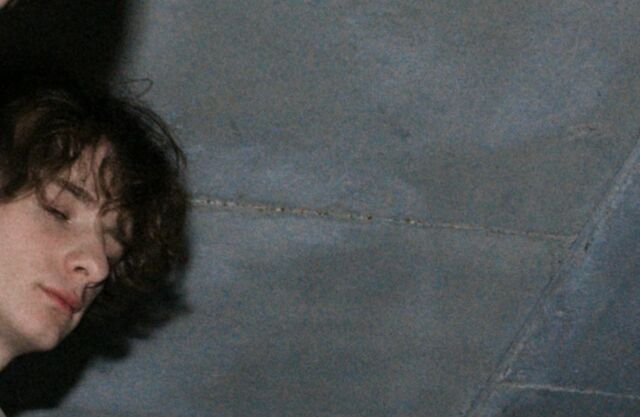 Close up of a young man with curly hair, eyes half closed against a grey wall