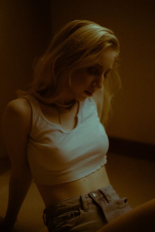 A woman in a white top leaning against a wall in dim warm light