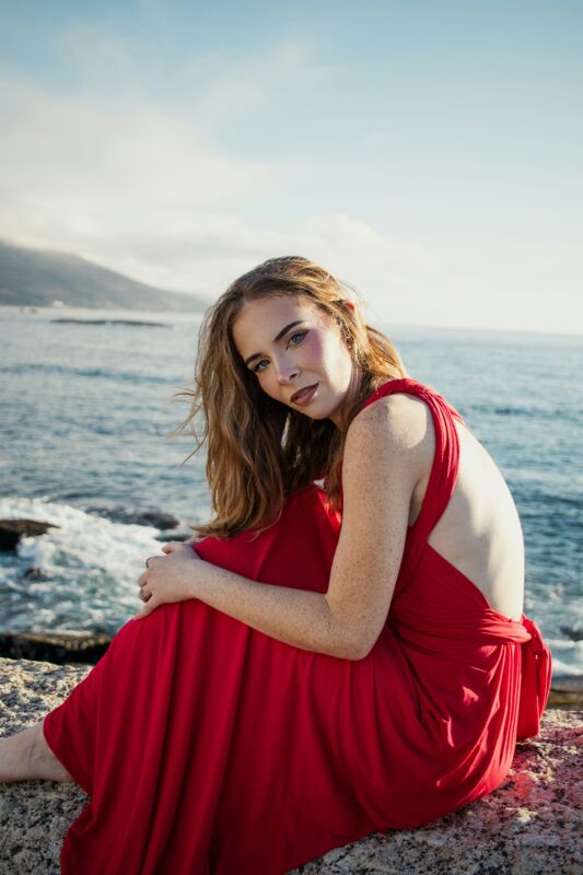 A woman in a red dress seated on rocks by the sea, looking at the camera