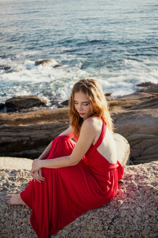 A woman in a red dress seated on rocks by the sea, looking away, a near duplicate frame