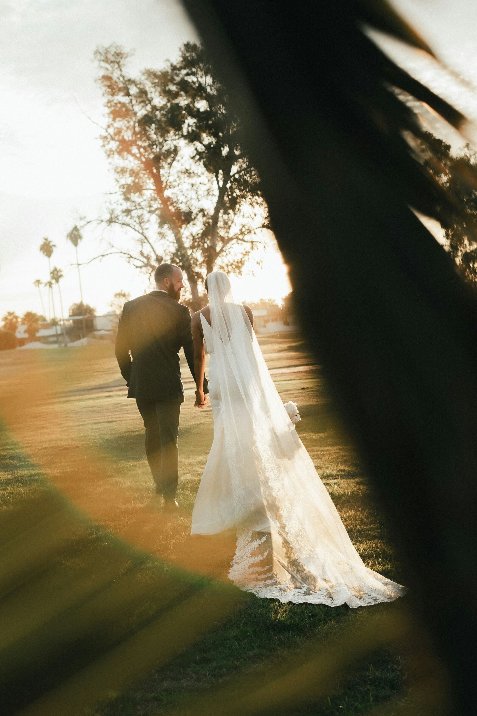 A bride and groom walking away across a lawn at golden hour, her veil catching the light